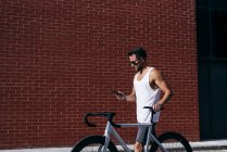 Vista lateral do belo ciclista masculino em sportswear e óculos de sol usando smartphone enquanto está de pé com bicicleta ao lado da parede de tijolo vermelho — Fotografia de Stock