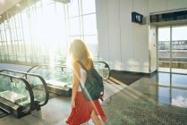 Vista lateral da encantadora mulher encaracolada em saia vermelha com mochila andando para baixo na escada rolante no aeroporto no Texas — Fotografia de Stock