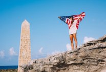 Fröhlich gebräunte Frau im Urlaub steht auf Klippen und hält amerikanische Flagge neben gerocktem Obelisk — Stockfoto