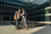 Fröhliche Männer und Frauen mit Fahrrad lächeln und schauen auf ein Tablet, während sie außerhalb des Bürogebäudes kommunizieren — Stockfoto