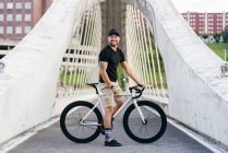 Счастливый взрослый бородатый мужчина в черной кепке в черной рубашке и бежевых шортах стоит с велосипедом через пешеходный мост в городе глядя в сторону — стоковое фото