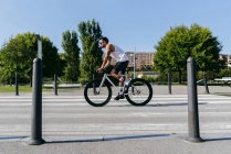 Side view of athletic man in sunglasses wearing white shirt and black shorts riding bike on city roadway with green trees on roadside on summer day with blue sky — Stock Photo