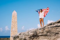Mujer bronceada alegre en vacaciones de pie en los acantilados y la celebración de la bandera americana por el obelisco balanceado - foto de stock