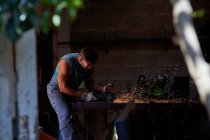 Views from tree branch leaves of workman in protective glasses and gloves cutting metal with grinder with flying sparks while working in workshop — Stock Photo
