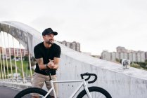 Счастливый взрослый бородатый мужчина в черной кепке в черной рубашке и бежевых шортах, стоящий на велосипеде, сидит через пешеходный мост в городе и смотрит в сторону — стоковое фото