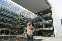 Glücklicher Mann trägt Frau huckepack beim Stehen und Lachen auf der Straße gegen zeitgenössisches Geschäftszentrum — Stockfoto