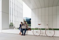 Zufriedene Geschäftsleute lächeln und stöbern gemeinsam am Laptop, während sie vor dem modernen Gebäude in der Nähe des Fahrrads auf der Stadtstraße sitzen — Stockfoto