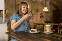 Блондинка весела щаслива жінка з чубчиком тримає смартфон над шматочком торта і сидить за столом з кавою і десертом — стокове фото