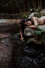 Довгошерста гола жінка з закритими очима лежить на камені і торкається води в таємничій річці в лісі — стокове фото