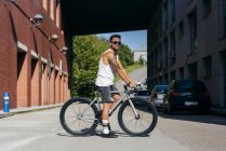 Sportive man in sunglasses wearing white sleeveless shirt and black shorts sitting on bicycle between buildings connected by bridge on summer sunny day — Stock Photo