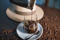 Gießen von aromatischen braunen Getränken im Glas auf einer runden Untertasse zwischen gerösteten Kaffeebohnen neben dem Hut auf einem glänzenden Tisch — Stockfoto