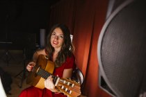 Charmante künstlerische Frau im roten Kleid, die bei warmem Licht auf der Bühne ein Lied auf der Gitarre spielt — Stockfoto