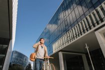 From below of pensive man making phone call while standing with bike against contemporary high rise business center — Stock Photo