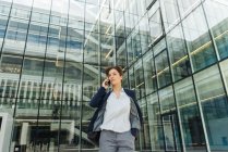 Angolo basso di felice donna d'affari che parla su smartphone mentre si trova fuori dal centro commerciale sulla strada della città — Foto stock