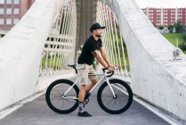 Щасливий дорослий бородатий чоловік у чорній шапці в чорній сорочці та бежевих шортах, що стоять з велосипедом через пішохідний міст у місті — стокове фото