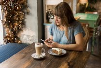 Блондинка молода весела жінка друкує повідомлення на смартфоні, сидячи за столом з кавою і десертом в кафе — стокове фото