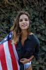 Allegro hipster donna dai capelli ricci con bandiera americana guardando in macchina fotografica da piante verdi — Foto stock