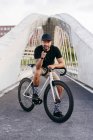 Щасливий дорослий бородатий чоловік у чорній шапці в чорній сорочці та бежевих шортах, що сидить на велосипеді через пішохідний міст у місті, дивлячись на камеру — стокове фото