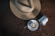 De arriba bebida marrón con espuma blanca en taza de cerámica entre granos de café al lado del sombrero y taza vacía en la mesa de madera - foto de stock