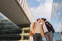 Внизу счастливая пара коллег целуют друг друга, стоя перед современным зданием на городской улице после работы — стоковое фото