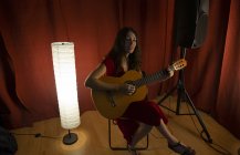 Чарівна художня жінка в червоній сукні виконує пісню, граючи на гітарі на сцені з теплим світлом — стокове фото