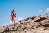 Cheerful woman wrapped in American flag standing on rocky cliffs against blue sky — Stock Photo