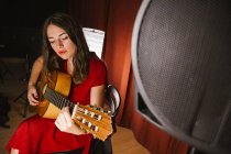 Affascinante donna artistica in abito rosso che suona la canzone sulla chitarra in scena con luce calda — Foto stock