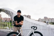 Счастливый взрослый бородатый мужчина в черной кепке в черной рубашке и бежевых шортах, стоящий на велосипеде, сидит через пешеходный мост в городе и смотрит в сторону — стоковое фото