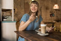 Blonde cheerful happy female with bangs holding spoon with piece of cake and sitting at table with coffee and dessert — Stock Photo