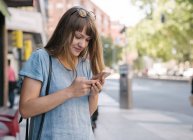 Blonde young cheerful female with sunglasses in blue T-shirt standing on street of city and typing message on smartphone — Stock Photo
