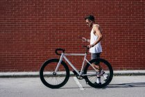 Side view of handsome male cyclist in sportswear and sunglasses using smartphone while standing with bike next to red brick wall — Stock Photo