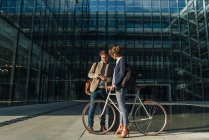 Homem e mulher alegre com bicicleta sorrindo e olhando um para o outro enquanto se comunica fora do prédio de escritórios — Fotografia de Stock