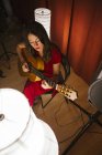 Dall'alto donna di talento in abito rosso eseguire canzone e suonare la chitarra in palcoscenico caldo illuminato vicino lampada bianca — Foto stock
