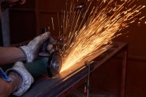 Cropped image of workman in protective glasses and gloves cutting metal with grinder with flying sparks while working in workshop — Stock Photo