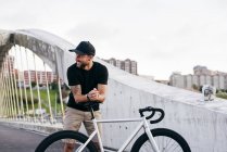 Hombre barbudo adulto feliz en gorra negra con camisa negra y pantalones cortos beige de pie apoyado en bicicleta sentarse a través de pasarela en la ciudad mirando hacia otro lado - foto de stock