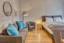 Quarto confortável com paredes de madeira branca e grande cama macia com sofá cinza aconchegante perto de lâmpada de assoalho ardente — Fotografia de Stock