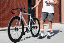 Обрезанный образ велосипедиста в спортивной одежде с помощью смартфона, стоя с велосипедом рядом с красной кирпичной стеной — стоковое фото