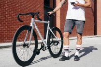 Ausgeschnittenes Bild eines Radfahrers in Sportbekleidung mit Smartphone, der mit Fahrrad neben roter Ziegelwand steht — Stockfoto