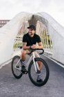 Homem barbudo adulto feliz em boné preto vestindo camisa preta e shorts bege sentado descansando na bicicleta através da passarela na cidade olhando para longe — Fotografia de Stock