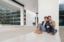 Весела пара обіймає один одного під час перегляду смартфона, сидячи за межами сучасної будівлі на міській вулиці разом — стокове фото