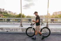Side view of young casual bearded cyclist in black cap using phone while walking with bike on road with city buildings on background — Stock Photo