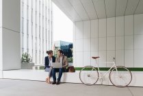 Задоволені бізнесмени посміхаються і переглядають ноутбук разом, сидячи за межами сучасної будівлі біля велосипеда на міській вулиці — стокове фото