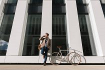 Baixo ângulo de empreendedor sério com smartphone apoiado na parede perto de uma bicicleta no edifício moderno e olhando para o centro da cidade — Fotografia de Stock