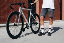 Image recadrée de cycliste en vêtements de sport à l'aide d'un smartphone tout en restant debout avec le vélo à côté du mur de briques rouges — Photo de stock