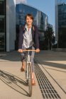 Delighted female entrepreneur smiling and riding bike on sunny day in downtown of modern city — Stock Photo
