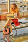 Стильная веселая женщина, печатающая на ноутбуке, сидя со скрещенными ногами на металлической скамейке в стеклянном коридоре аэропорта в Техасе — стоковое фото