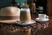 Verre de boisson chaude en composition avec chapeau et grains de café sur table en bois — Photo de stock