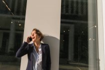 Ottimista donna d'affari in giacca scura sorridente e distogliendo lo sguardo mentre si appoggia sul muro di costruzione sulla strada della città e risponde alla telefonata — Foto stock