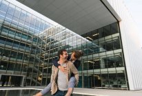 Happy adult man in casual clothes carry woman piggyback while standing and laughing on street against contemporary business center in downtown — Stock Photo