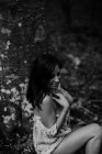 Vista lateral de una mujer encantadora y de pelo largo tocando la piel a mano y sentado en piedra en un misterioso bosque. - foto de stock